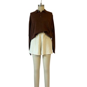 May include: A brown long-sleeve polo shirt with a three-button placket is paired with off-white shorts and beige leggings. The outfit is displayed on a mannequin against a white background. The leggings have a decorative trim at the ankles.