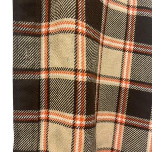 May include: Close-up of a brown, tan, and orange plaid fabric. The plaid pattern features large squares with orange and white accent lines. The fabric has a textured appearance and a button is visible at the top.