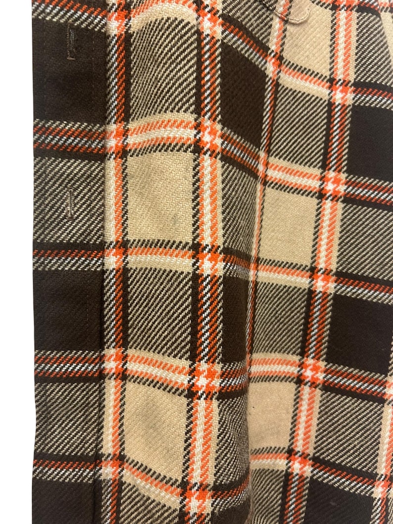May include: Close-up of a brown, tan, and orange plaid fabric. The plaid pattern features large squares with orange and white accent lines. The fabric has a textured appearance and a button is visible at the top.