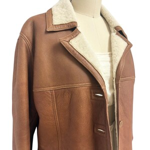 May include: A brown leather jacket with a cream-colored shearling lining and collar. The jacket has a classic design with a button-up front and two front pockets. The jacket is displayed on a mannequin.
