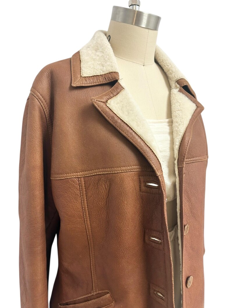 May include: A brown leather jacket with a cream-colored shearling lining and collar. The jacket has a classic design with a button-up front and two front pockets. The jacket is displayed on a mannequin.