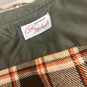 May include: Close-up of a vintage garment with a plaid pattern in brown, beige, and orange. The interior lining is olive green. A label reads "DESIGNED BY Carl Michaels" in red script.