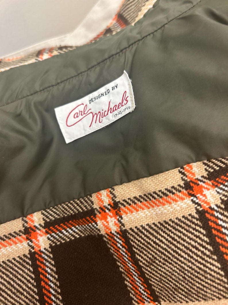 May include: Close-up of a vintage garment with a plaid pattern in brown, beige, and orange. The interior lining is olive green. A label reads "DESIGNED BY Carl Michaels" in red script.