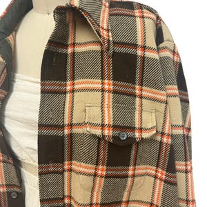 May include: A brown, tan, and orange plaid shirt with two front pockets and a button closure. The shirt is displayed on a mannequin. The collar and inner lining are a darker shade of brown.