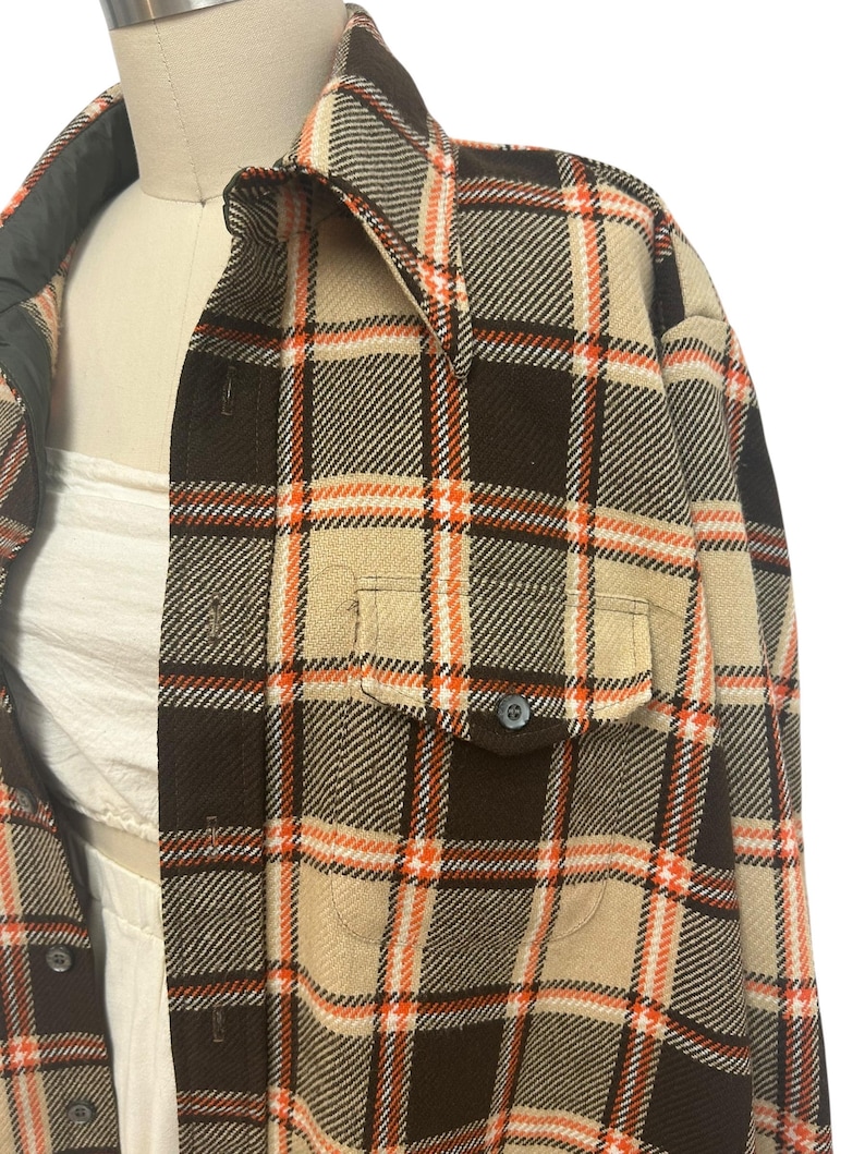 May include: A brown, tan, and orange plaid shirt with two front pockets and a button closure. The shirt is displayed on a mannequin. The collar and inner lining are a darker shade of brown.