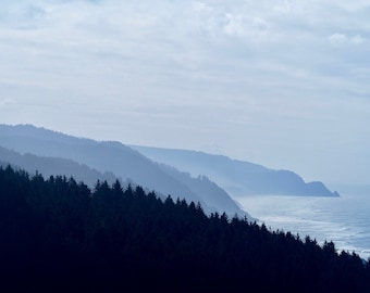 May include: A scenic landscape photograph of a coastal mountain range. The image features layers of blue-toned mountains, a dark green forest, and the ocean under a cloudy sky. The scene evokes a sense of tranquility and natural beauty.