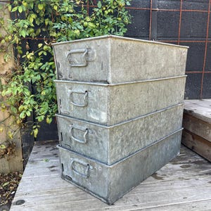 May include: Four rectangular galvanized metal storage bins stacked on top of each other. The bins have handles and a weathered, muted grey appearance. They sit on a wooden deck, with greenery and a dark fence visible in the background.