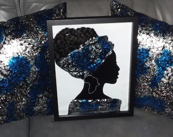 Könnte beinhalten: Eine gerahmte Silhouette einer Frau mit einem blauen und silbernen Pailletten-Kopftuch und einem großen Ohrring in Form des afrikanischen Kontinents. Der Rahmen ist schwarz und der Hintergrund ist weiß. Das Bild ist von zwei blauen und silbernen Pailletten-Kissen umgeben.