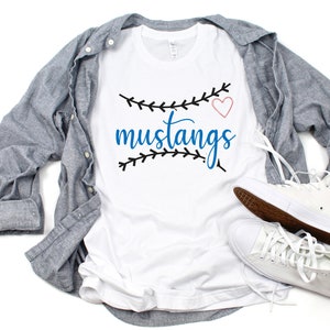 May include: White t-shirt with a blue and black baseball-themed design that says "mustangs" with a pink heart. The shirt is laid out on a white surface with a blue denim shirt and white high-top sneakers.