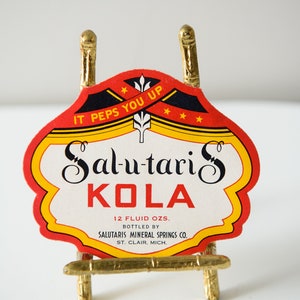 Könnte beinhalten: Ein Vintage-Etikett für Salutaris Kola, eine 355 ml-Flasche Limonade. Das Etikett ist rot, gelb und schwarz mit dem Text "IT PEPS YOU UP" und "Salutaris KOLA". Das Etikett befindet sich auf einem kleinen goldenen Staffelei.