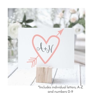 May include: A pink heart with an arrow through it, with the letters A+N inside the heart. The heart is on a white card, which is on a wooden block.