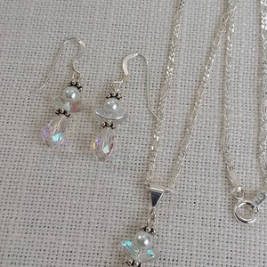 May include: A silver necklace and earrings set featuring crystal angel pendants with pearl accents. The necklace has a delicate silver chain.
