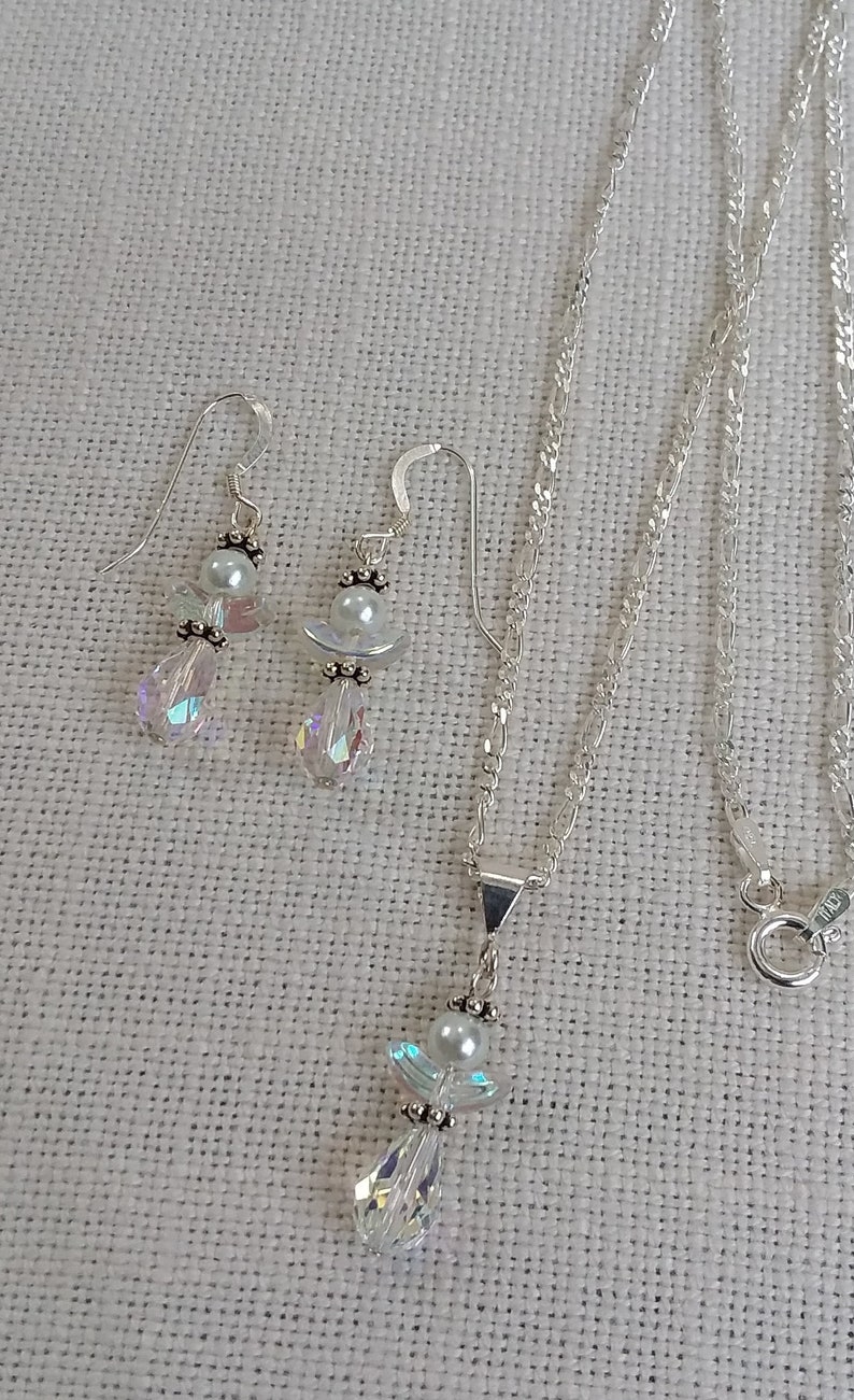 May include: A silver necklace and earrings set featuring crystal angel pendants with pearl accents. The necklace has a delicate silver chain.