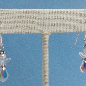 May include: A pair of silver dangle earrings featuring a crystal angel charm with a white pearl bead. The angel has iridescent wings and a teardrop-shaped crystal pendant.