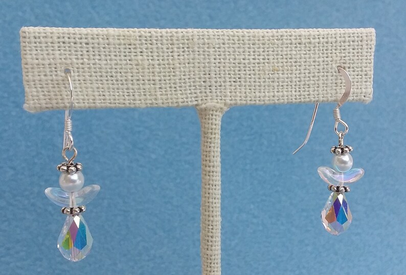 May include: A pair of silver dangle earrings featuring a crystal angel charm with a white pearl bead. The angel has iridescent wings and a teardrop-shaped crystal pendant.