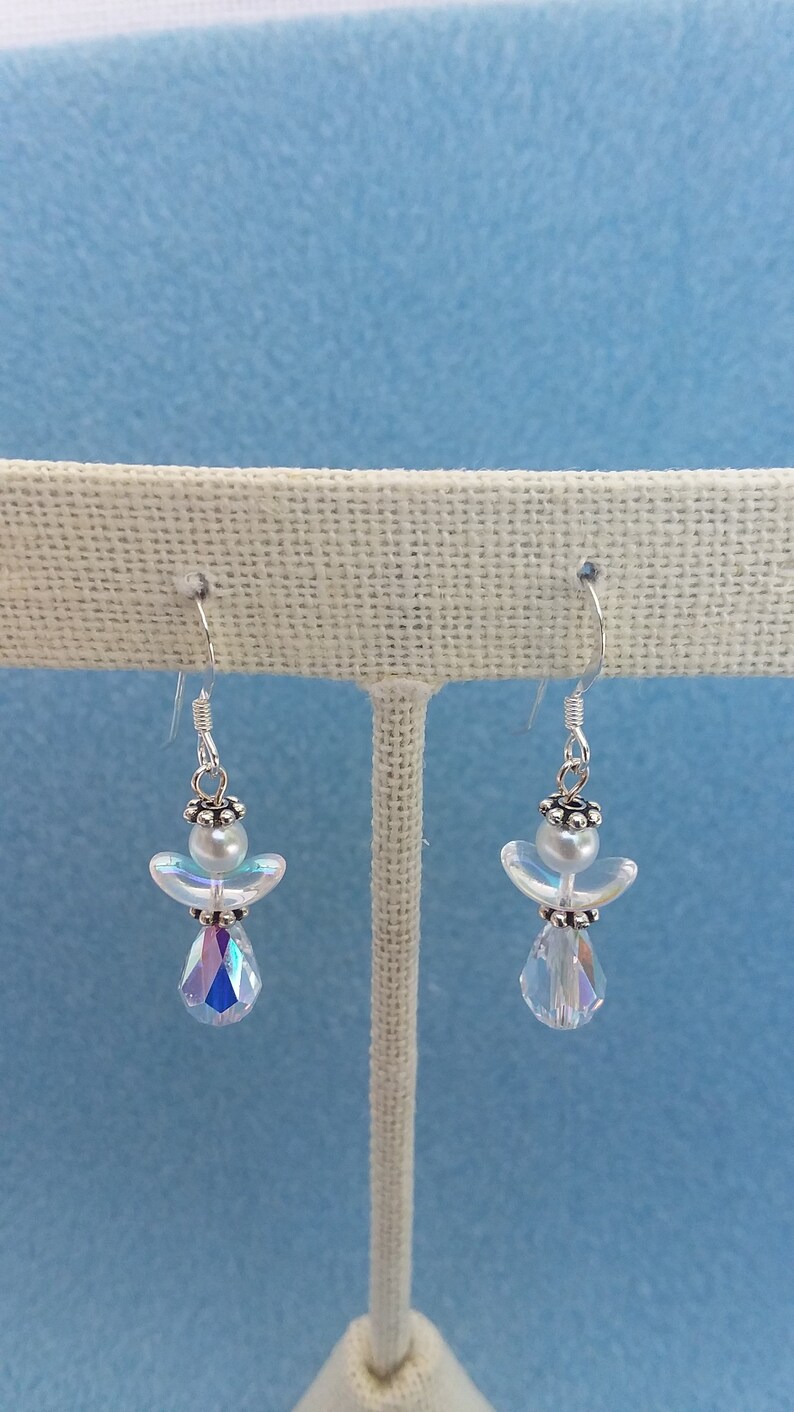 May include: A pair of silver angel dangle earrings with iridescent crystal drops. The angels are made of white beads and have silver wings.