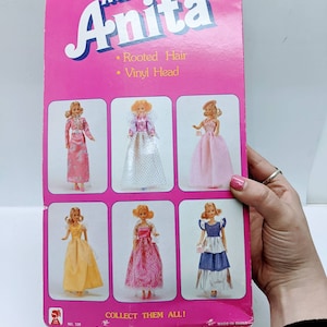 Pu&ograve; includere: Una scatola di cartone rosa e bianca con le parole "Miss Anita" in grandi lettere rosa. La scatola presenta un'immagine di sei bambole che indossano abiti diversi. Il testo sulla scatola recita "Capelli radicati" e "Testa in vinile". La scatola dice anche "Collezionali tutti!" e "N. 128".