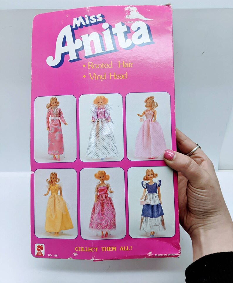 Pu&ograve; includere: Una scatola di cartone rosa e bianca con le parole "Miss Anita" in grandi lettere rosa. La scatola presenta un'immagine di sei bambole che indossano abiti diversi. Il testo sulla scatola recita "Capelli radicati" e "Testa in vinile". La scatola dice anche "Collezionali tutti!" e "N. 128".