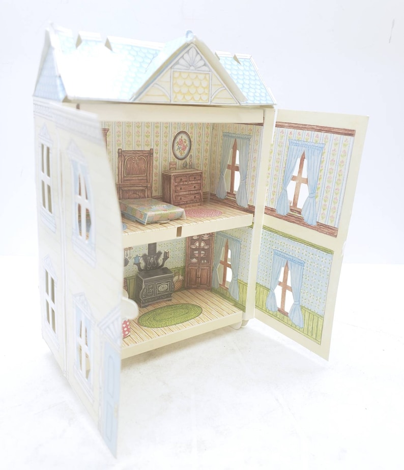 Puede incluir: Una casa de mu&ntilde;ecas de cart&oacute;n con dos habitaciones, un dormitorio y una cocina. La casa de mu&ntilde;ecas est&aacute; pintada de blanco con molduras azules y tiene un techo azul claro. La casa de mu&ntilde;ecas est&aacute; abierta y el interior est&aacute; decorado con papel tapiz y muebles.