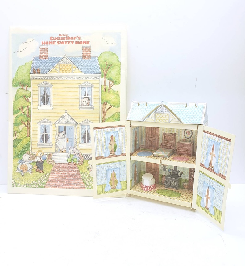 Puede incluir: Una casa de mu&ntilde;ecas de papel amarilla y azul con las palabras "Missy Cucumber's Home Sweet Home" en la parte delantera. La casa de mu&ntilde;ecas est&aacute; abierta y muestra el interior de la casa, que est&aacute; decorada con muebles y papel tapiz.