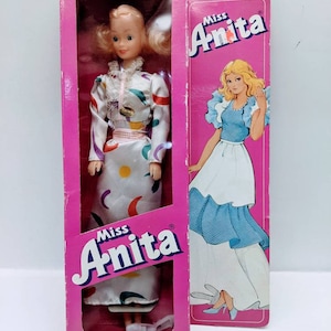 Pu&ograve; includere: Una bambola vintage Miss Anita con un vestito bianco con motivi geometrici colorati. La bambola &egrave; confezionata in una scatola rosa con il logo Miss Anita. La scatola presenta anche un'immagine della bambola con un vestito blu.