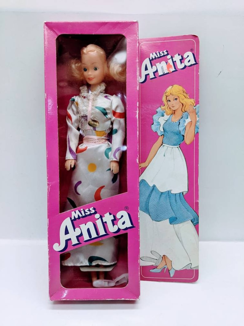 Pu&ograve; includere: Una bambola vintage Miss Anita con un vestito bianco con motivi geometrici colorati. La bambola &egrave; confezionata in una scatola rosa con il logo Miss Anita. La scatola presenta anche un'immagine della bambola con un vestito blu.