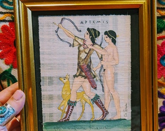 May include: A framed print on papyrus depicting Artemis, the Greek goddess of the hunt, with a bow and arrow, accompanied by a dog. The print is signed "De Fiori" and includes the text "Artemis, goddess of the hunt".