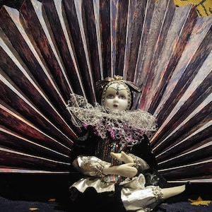 May include: A porcelain doll with a harlequin costume, featuring a black velvet jacket, silver accents, and a ruffled collar. The doll is seated in front of a fan-shaped backdrop with brown and white stripes. The doll has a painted face and is wearing a jeweled headdress.