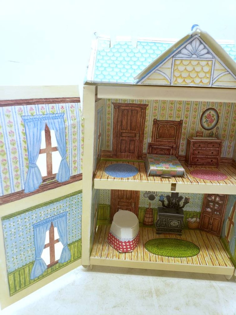 Puede incluir: Una casa de mu&ntilde;ecas de cart&oacute;n vintage con un techo azul y un exterior blanco. La casa de mu&ntilde;ecas tiene dos pisos y est&aacute; abierta para revelar las habitaciones interiores. Las habitaciones interiores est&aacute;n decoradas con papel tapiz y muebles. La casa de mu&ntilde;ecas es un gran juguete para ni&ntilde;os.