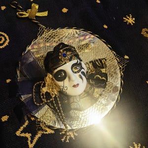 May include: A decorative mask ornament with a white face, black and gold accents, and a blue jewel. The mask has a painted design and is surrounded by gold netting and a blue ribbon. A gold bow and string are attached for hanging.