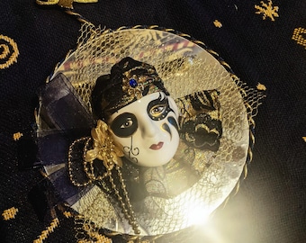 May include: A decorative mask ornament with a white face, black and gold accents, and a blue jewel. The mask has a painted design and is surrounded by gold netting and a blue ribbon. A gold bow and string are attached for hanging.