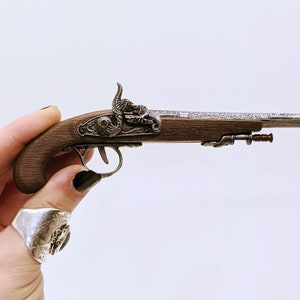 May include: A small, silver and brown, decorative, antique-style pistol with intricate details. The pistol is held in a hand with a silver ring.