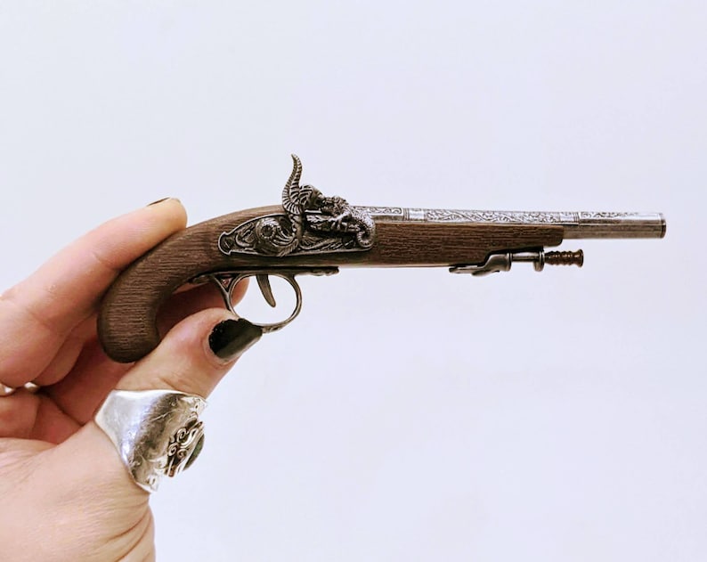 May include: A small, silver and brown, decorative, antique-style pistol with intricate details. The pistol is held in a hand with a silver ring.