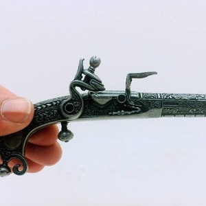 May include: A small, intricately detailed, black metal pistol with a green gemstone on the handle. The pistol is held in a hand wearing a silver ring with a green gemstone.