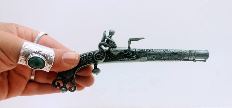 May include: A small, intricately detailed, black metal pistol with a green gemstone on the handle. The pistol is held in a hand wearing a silver ring with a green gemstone.