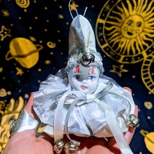 May include: A small porcelain jester doll in white and silver. The doll has a painted face with blue eyes and red accents, and a silver bell on its hat. The doll is held in a hand.
