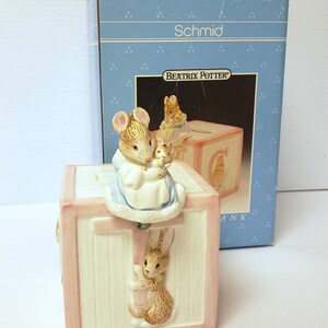 May include: A pink ceramic piggy bank shaped like a block with the letter T on it. The bank features a Beatrix Potter character, a mouse, holding a baby mouse. A rabbit is climbing the side of the block. The box says "Schmid" and "Beatrix Potter".