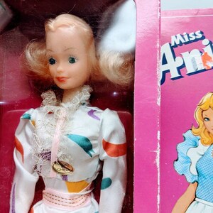 Pu&ograve; includere: Una bambola vintage Miss Annie con capelli biondi e un vestito bianco con motivi geometrici colorati. La bambola indossa una cintura rosa e un colletto di pizzo bianco. La bambola &egrave; confezionata in una scatola rosa con le parole "Miss Annie" stampate sul lato.
