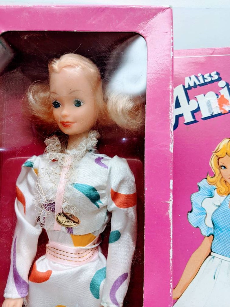 Pu&ograve; includere: Una bambola vintage Miss Annie con capelli biondi e un vestito bianco con motivi geometrici colorati. La bambola indossa una cintura rosa e un colletto di pizzo bianco. La bambola &egrave; confezionata in una scatola rosa con le parole "Miss Annie" stampate sul lato.