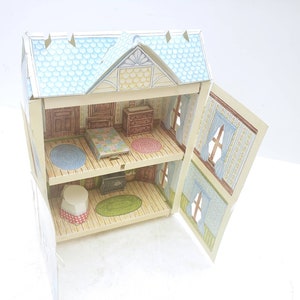 Puede incluir: Una casa de mu&ntilde;ecas de cart&oacute;n con dos niveles, que presenta un techo azul, una puerta delantera amarilla y un interior estampado. La casa de mu&ntilde;ecas est&aacute; abierta, revelando un dormitorio con una cama, una c&oacute;moda y una alfombra, y una sala de estar con una chimenea, una alfombra y una mesa.