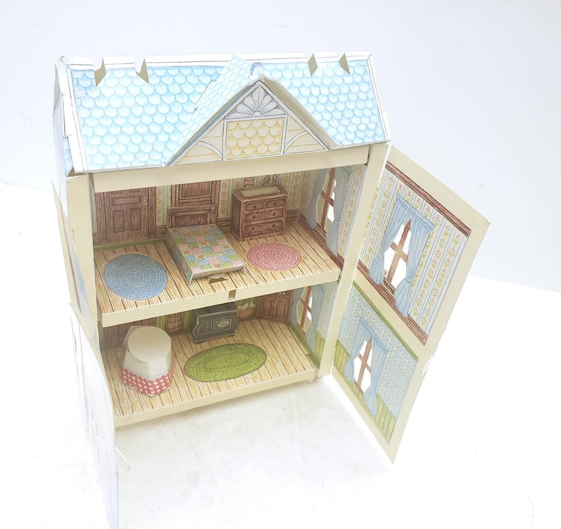 Puede incluir: Una casa de mu&ntilde;ecas de cart&oacute;n con dos niveles, que presenta un techo azul, una puerta delantera amarilla y un interior estampado. La casa de mu&ntilde;ecas est&aacute; abierta, revelando un dormitorio con una cama, una c&oacute;moda y una alfombra, y una sala de estar con una chimenea, una alfombra y una mesa.