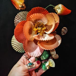 May include: A flower made of seashells with a turquoise ring on a hand. The flower is made of many different types of shells, including scallop shells, oyster shells, and conch shells. The flower is arranged in a circular pattern, with the turquoise ring in the center.