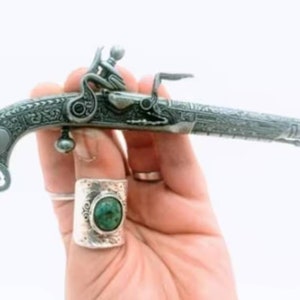 May include: A silver-colored, intricately engraved, miniature pistol with a black handle. The pistol is held in a hand wearing a silver ring with a green gemstone.