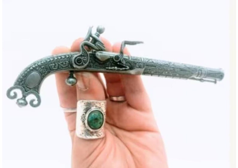 May include: A silver-colored, intricately engraved, miniature pistol with a black handle. The pistol is held in a hand wearing a silver ring with a green gemstone.