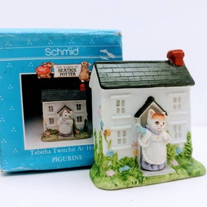 Puede incluir: Una figurita de cerámica blanca de Tabitha Twitchit del mundo de Beatrix Potter. La figurita representa a Tabitha de pie en la puerta de su casa, sosteniendo una bolsa azul y blanca a rayas. La casa tiene un techo verde y una chimenea roja.