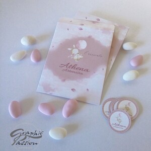 Può includere: Sacchetti regalo rosa e bianchi e adesivi abbinati con un'illustrazione di coniglietto, il nome "Athena Artemisia" e la parola "Essuata". Mandorle zuccherate bianche e rosa sparse.