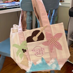 May include: A light pink and white striped canvas tote bag featuring the embroidered text Trader Joe's in a circular logo. The bag is decorated with fabric appliques including a green palm tree, a brown guitar, a pink flower, and blue waves.