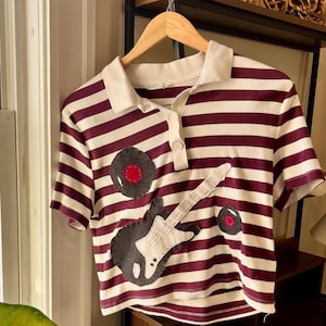 May include: A short-sleeved polo shirt with burgundy and white horizontal stripes. The shirt features a white collar and a button-up placket. Appliqued on the front are a black and white guitar and two vinyl record designs with red centers.