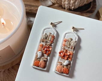 May include: A pair of rectangular dangle earrings featuring a white arched frame filled with miniature clay details including stacked books, orange flowers with small clear centers, small white ghosts, and orange pumpkins, attached to gold-tone posts.