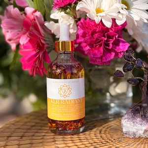 May include: A clear glass bottle with a gold cap and dropper, filled with a golden liquid and flower petals. The label reads "KLR RADIANT" and "Karma Light Readings." The bottle is in front of a bouquet of pink and white flowers.